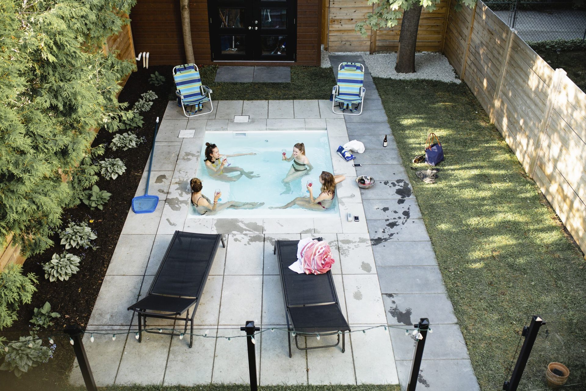 Four women relax in a small rectangular plunge pool in a fenced backyard, surrounded by lounge chairs, string lights, and shaded by mature trees and a wooden privacy fence.