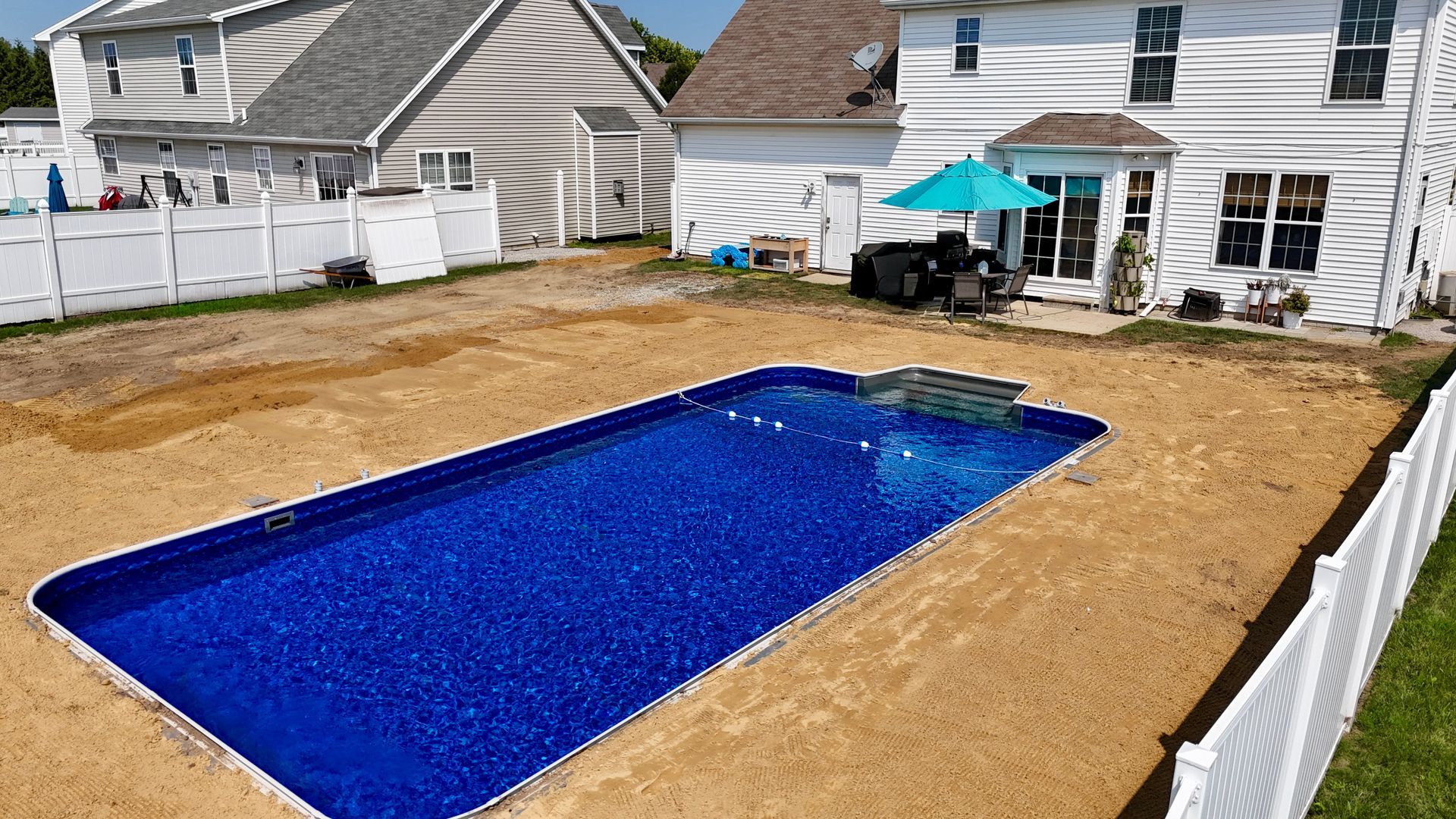 Rectangular blue swimming pool in a backyard with a white fence, grass, and a white house.