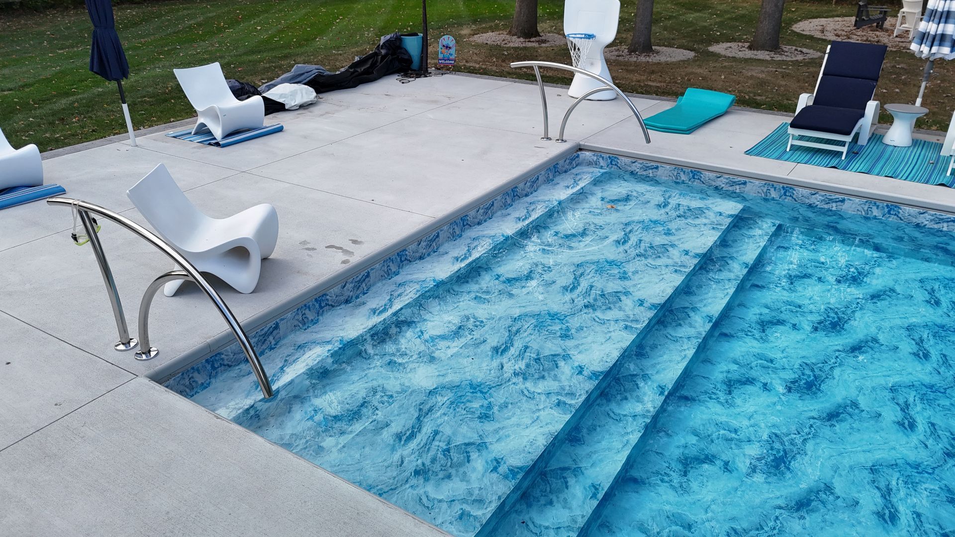 Swimming pool with steps, handrail, and lounge chairs on the concrete deck. Blue water and pool liner.