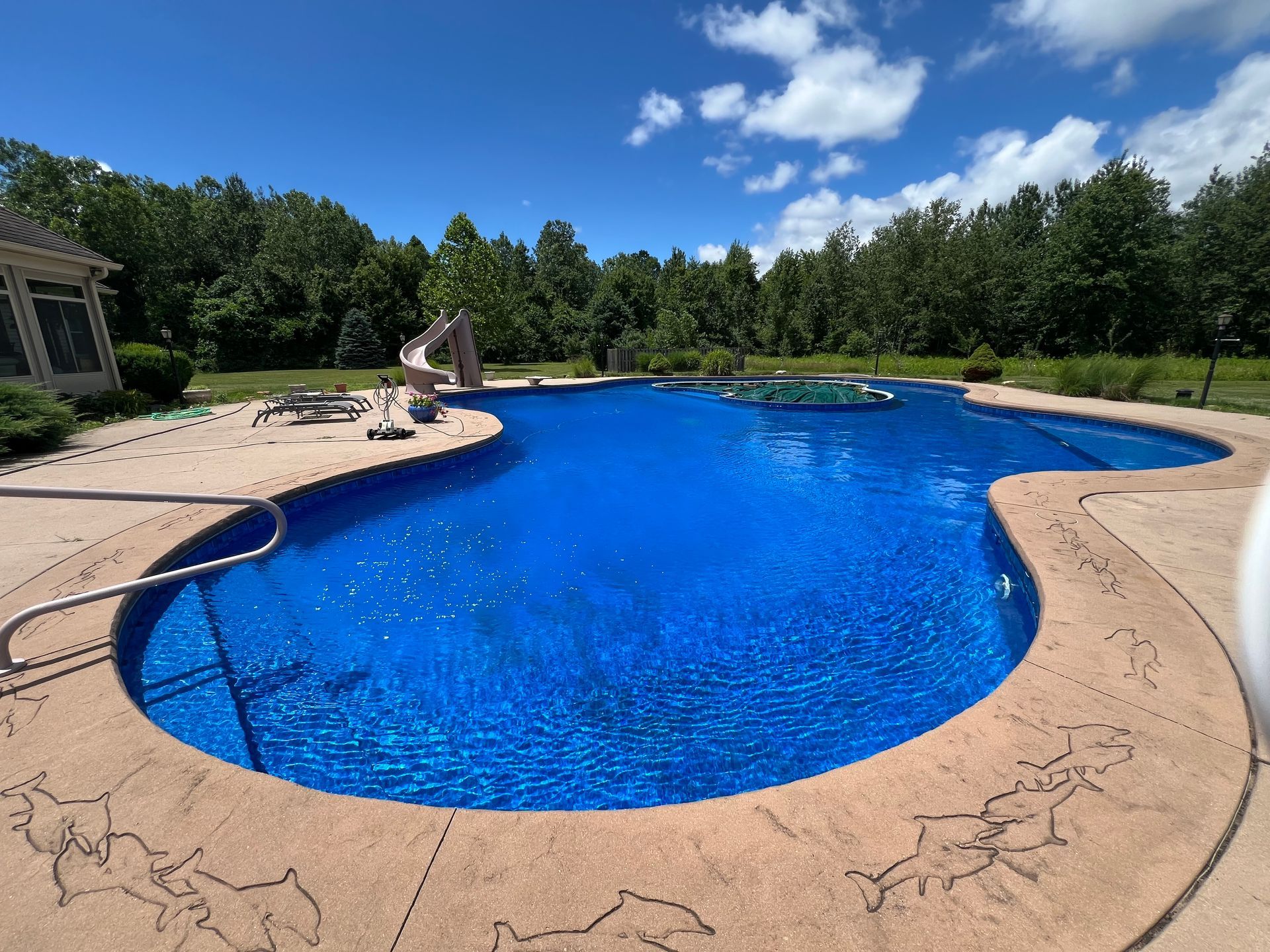 A large, blue swimming pool on a sunny day, with a slide and a surrounding concrete deck.