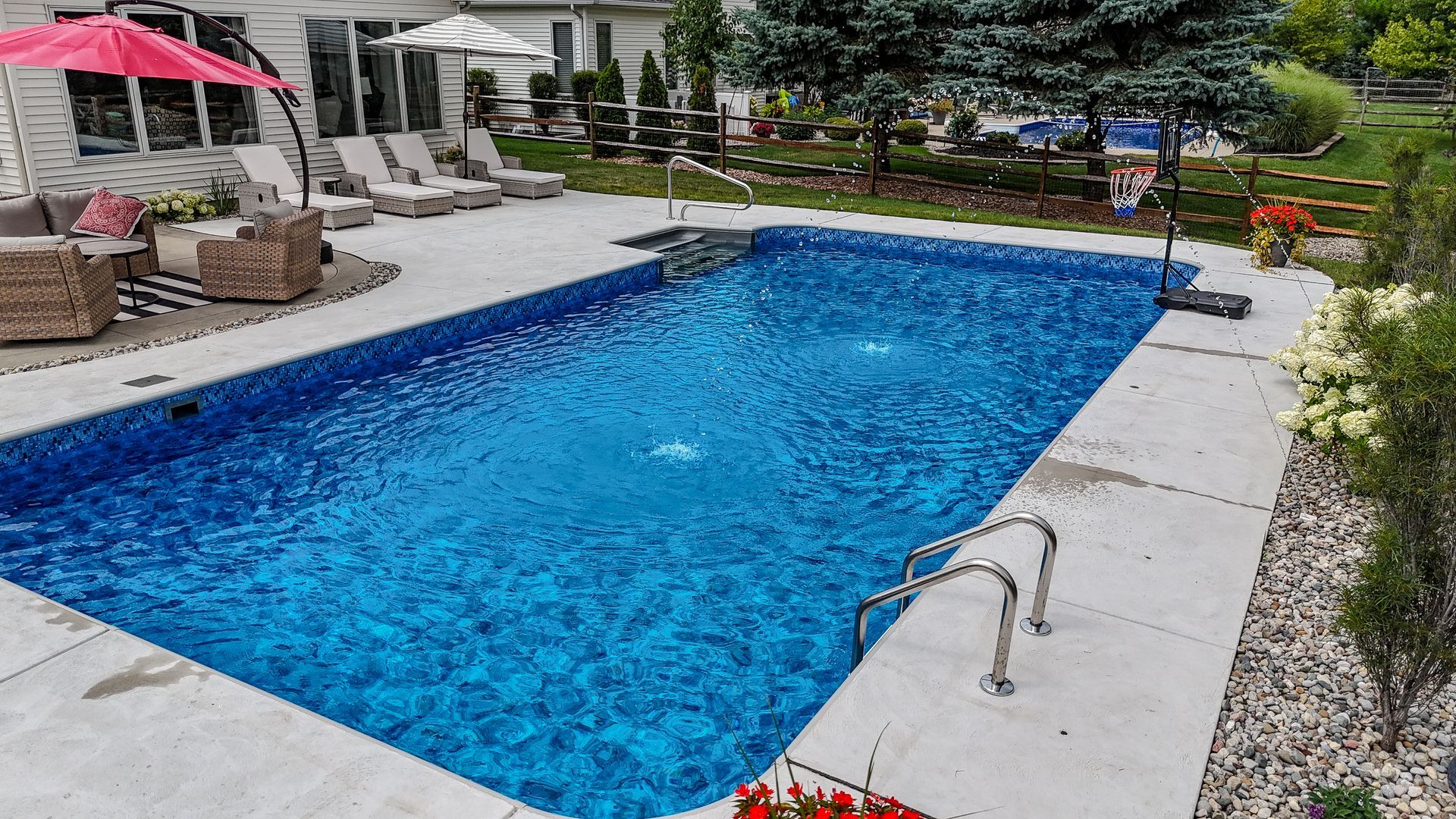 Rectangular blue-tiled pool with fountains, concrete deck, lounge chairs, and umbrella in a backyard setting.