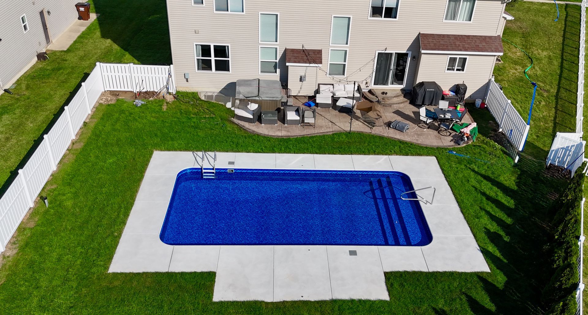 Overhead view of a rectangular blue swimming pool in a backyard with a house, patio, and green grass.