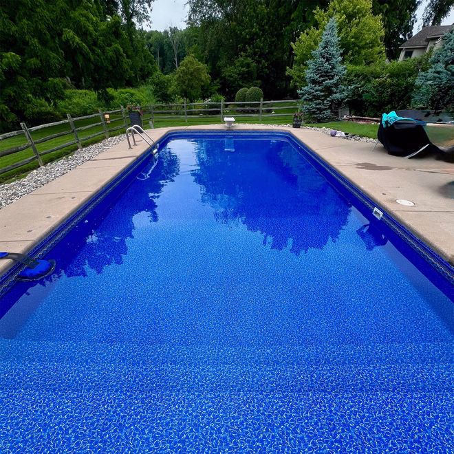 Rectangular blue pool surrounded by concrete, in a backyard setting with trees and a fence.