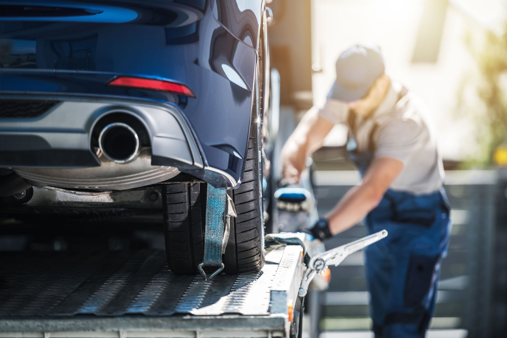 A worker in uniform secures a blue car onto the bed of a flatbed tow truck.