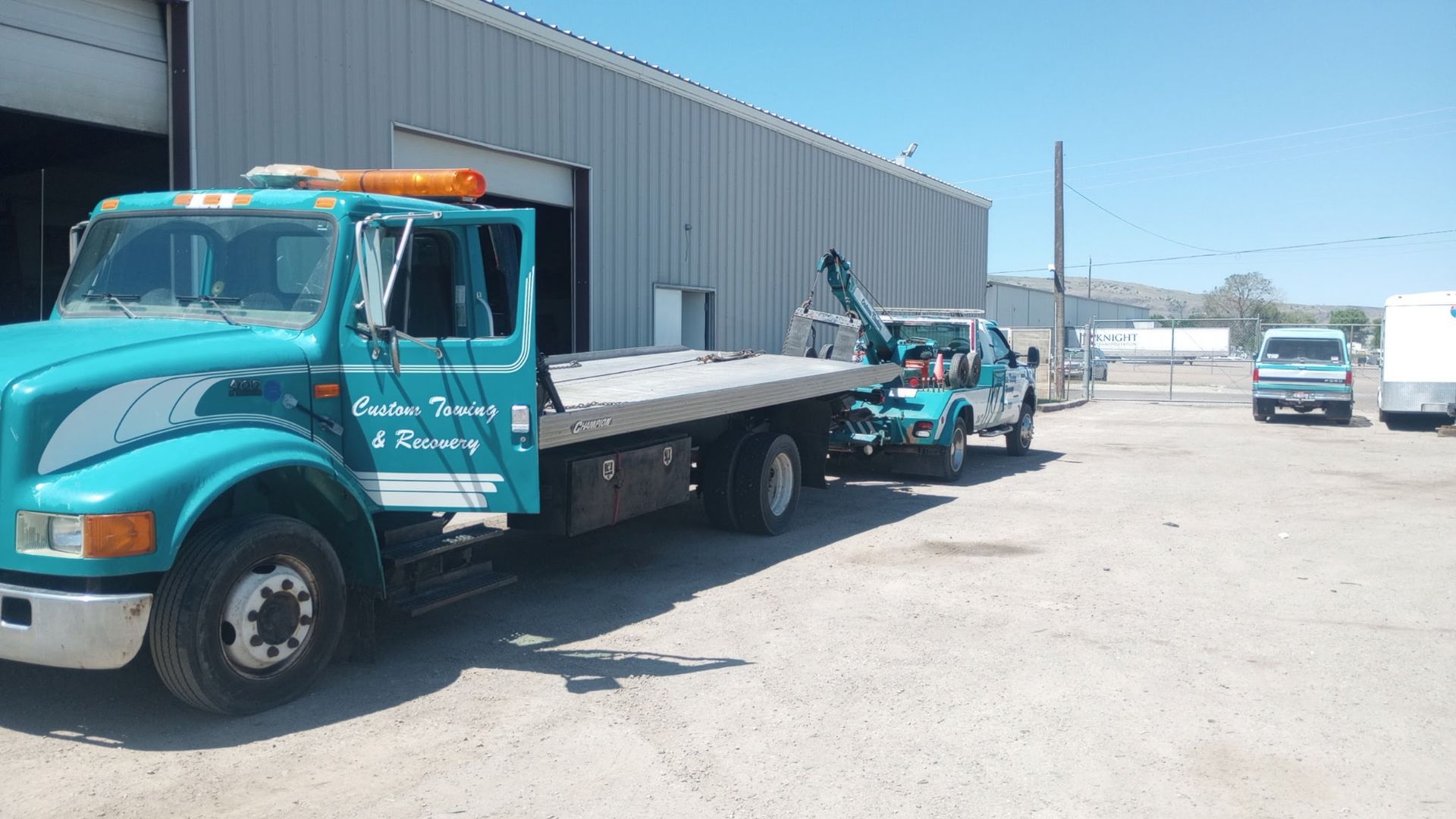 A teal flatbed tow truck parked outside a warehouse building on a sunny day.