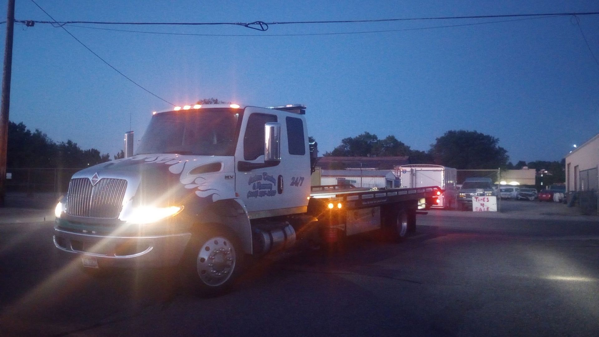 A white flatbed tow truck with its headlights and hazard lights on, parked in a lot during twilight.