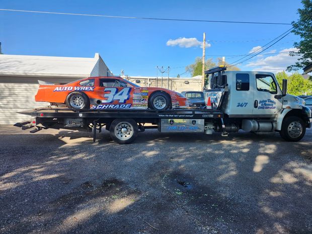 Tow truck hauling a red race car on a flatbed, parked on a sunny street under a blue sky.
