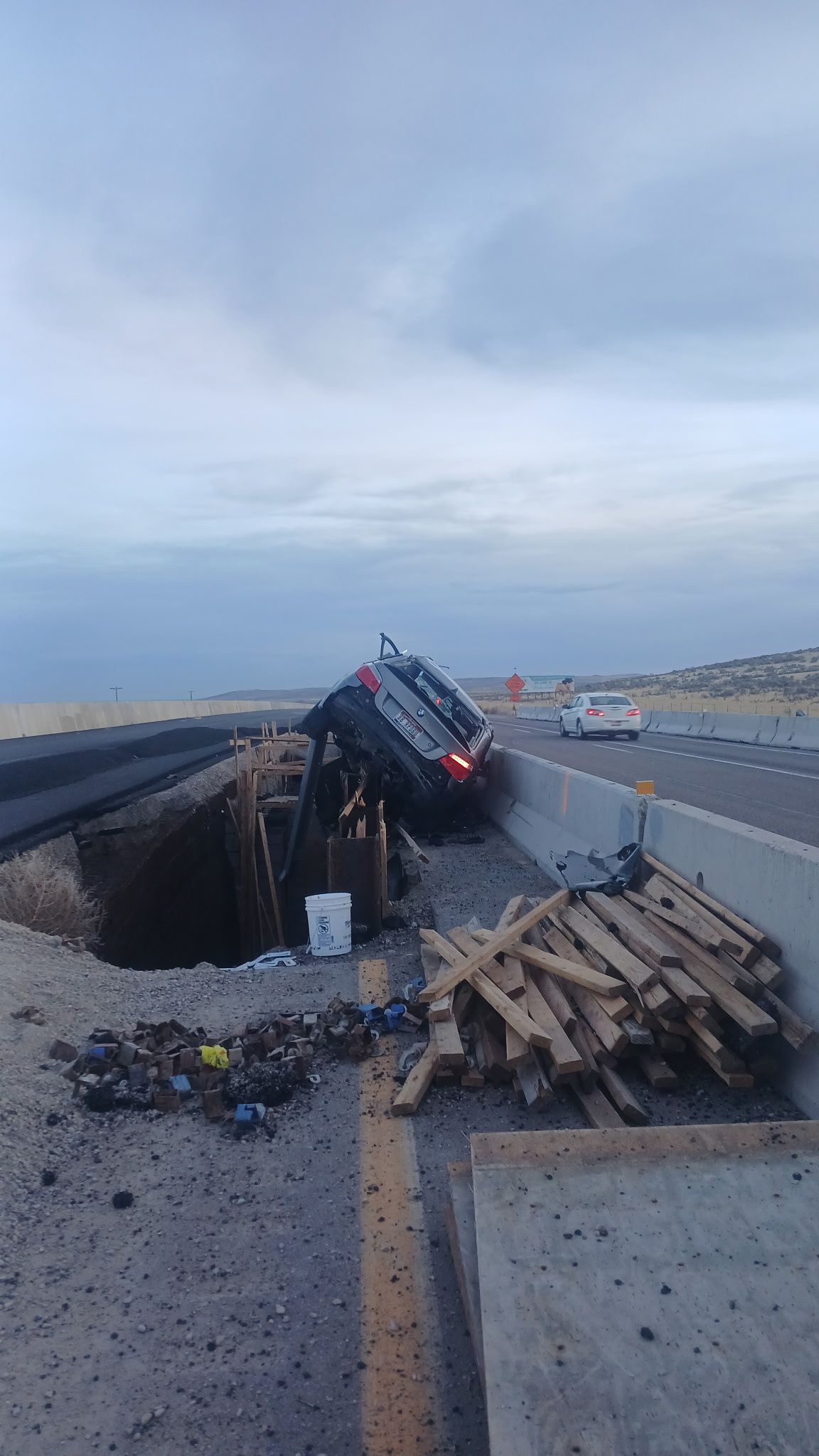 A car is teetering on the edge of a large construction pit on the side of a highway, with wooden planks nearby.