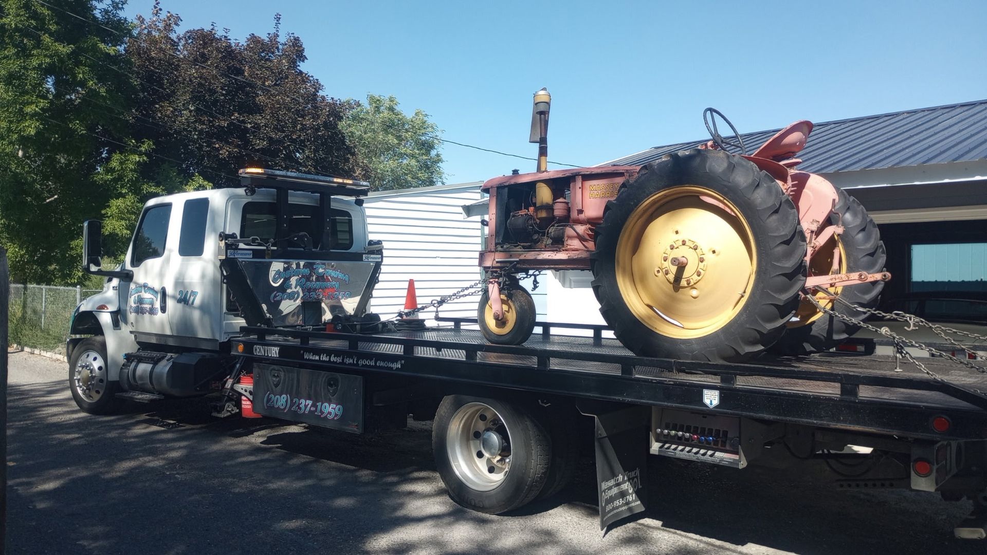 A white flatbed tow truck carries a vintage, rusty red and yellow farm tractor on a sunny day.