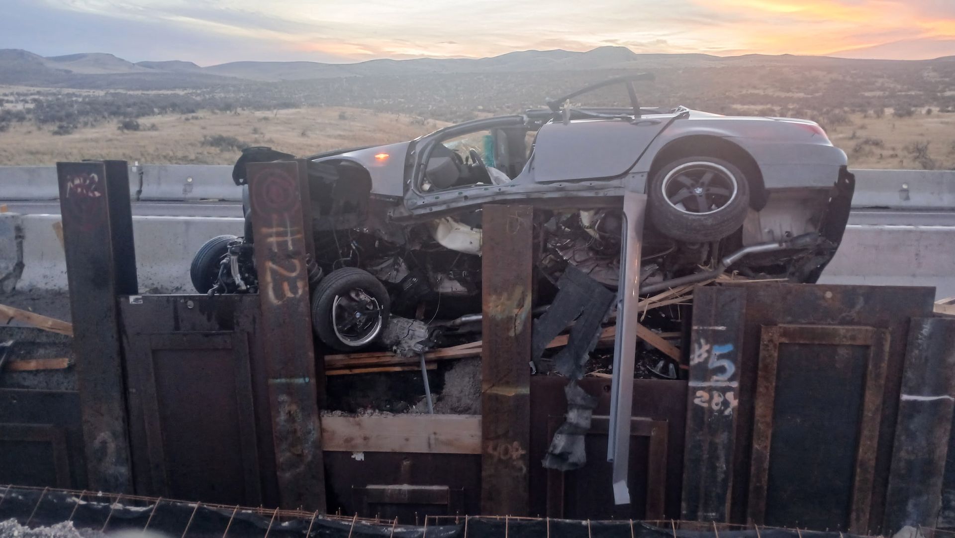 A severely damaged silver sedan crushed vertically against upright metal pillars on a highway at sunset.