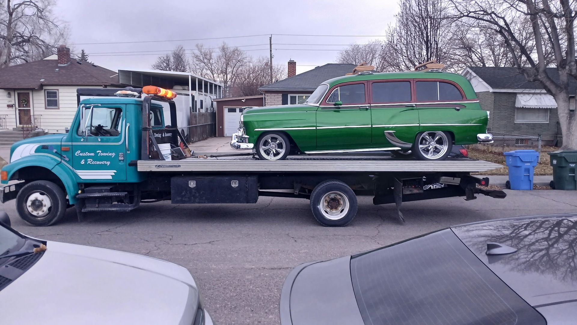 A teal flatbed tow truck carries a vintage, bright green station wagon on a residential street.