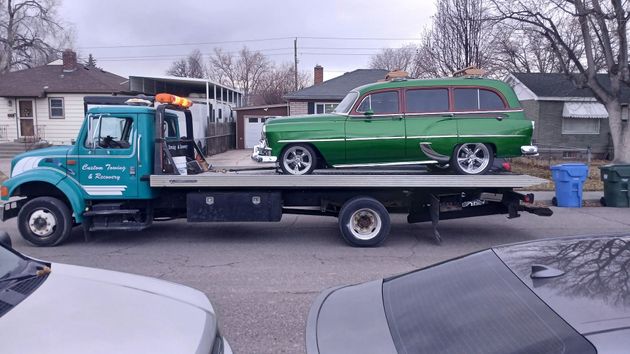 A green station wagon is parked on the flatbed of a teal tow truck on a residential street.