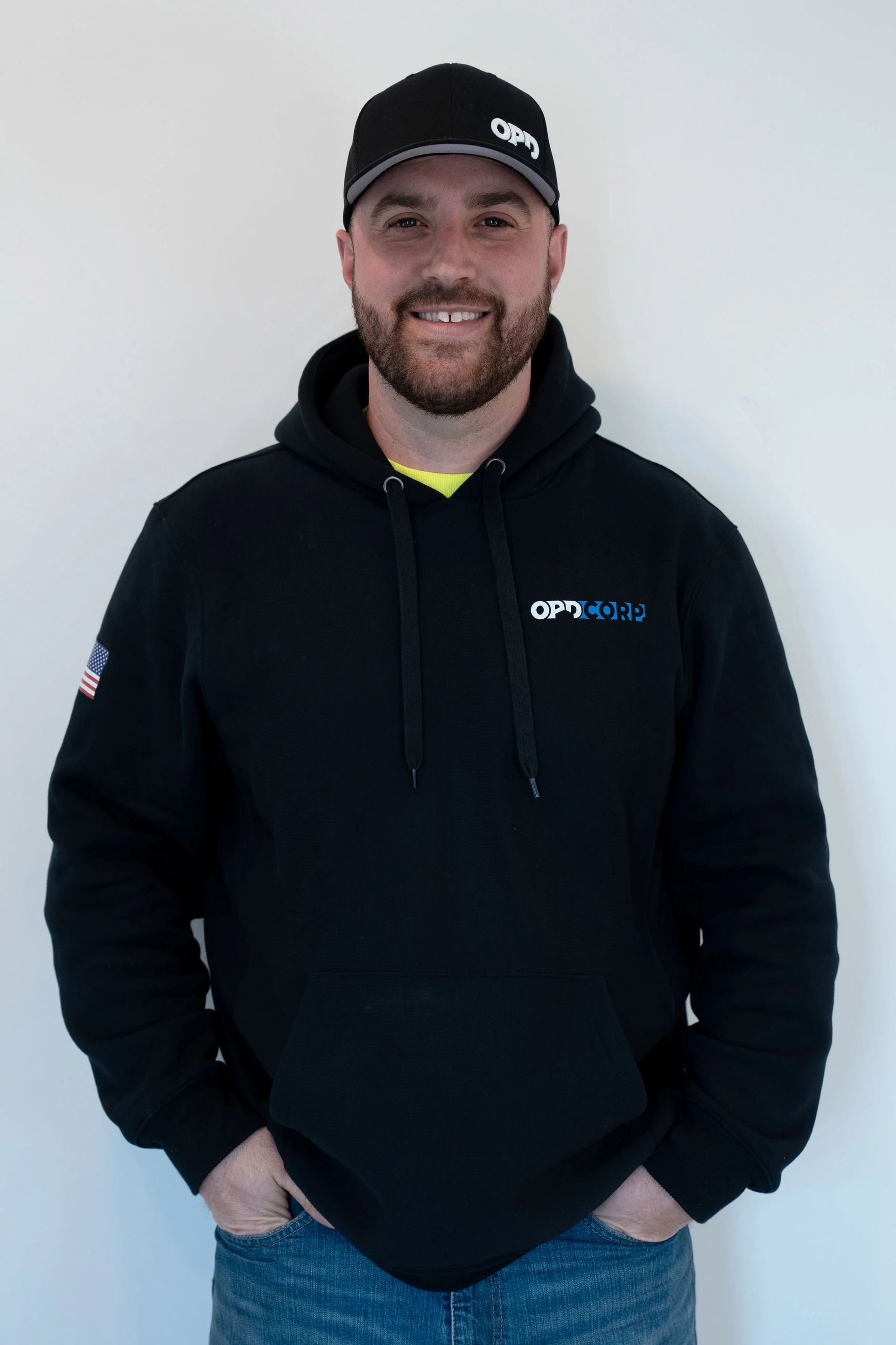 Man in black hoodie and hat, hands in pockets, smiling. White wall background.