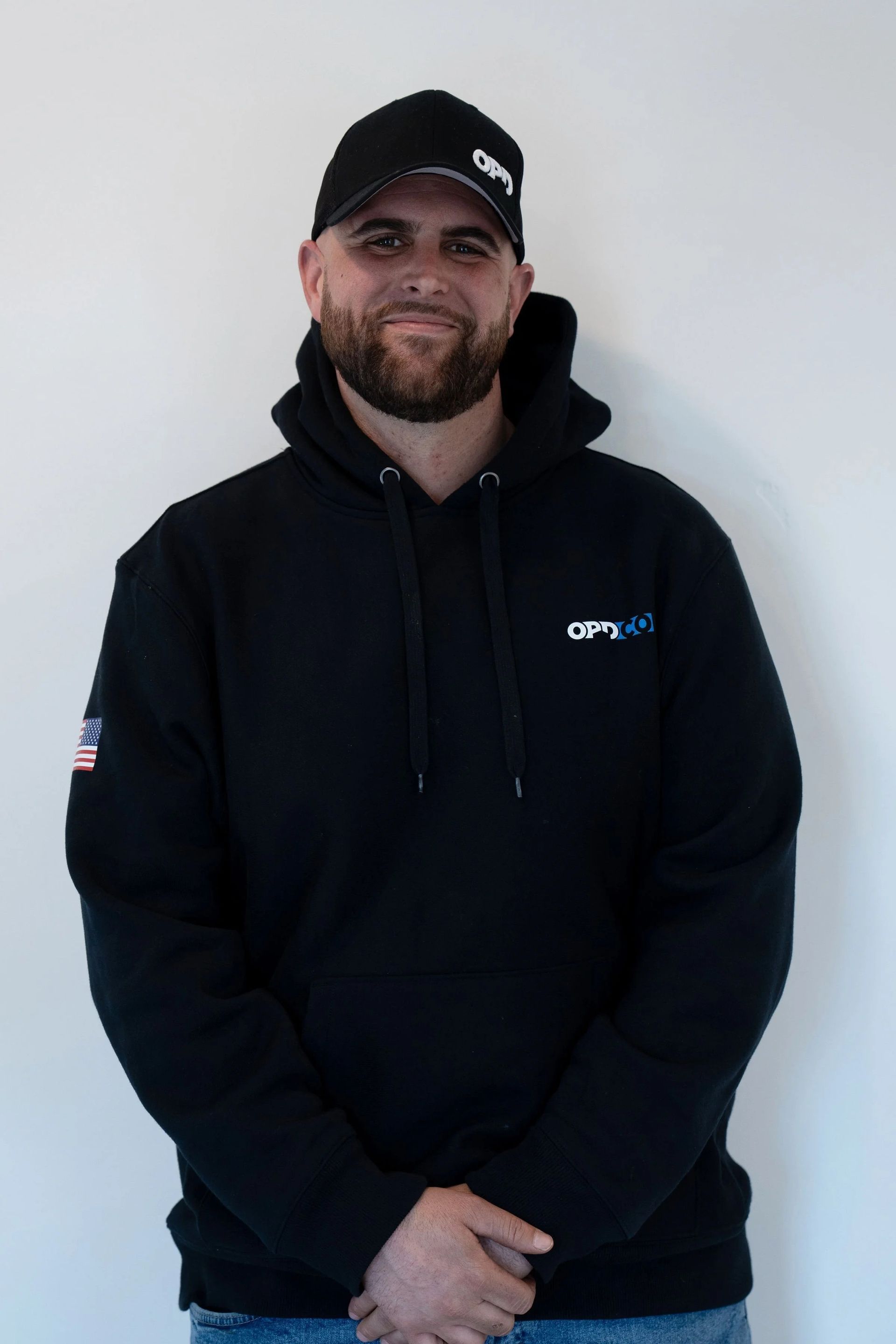 Man wearing a black hoodie and cap smiles, standing against a white wall.