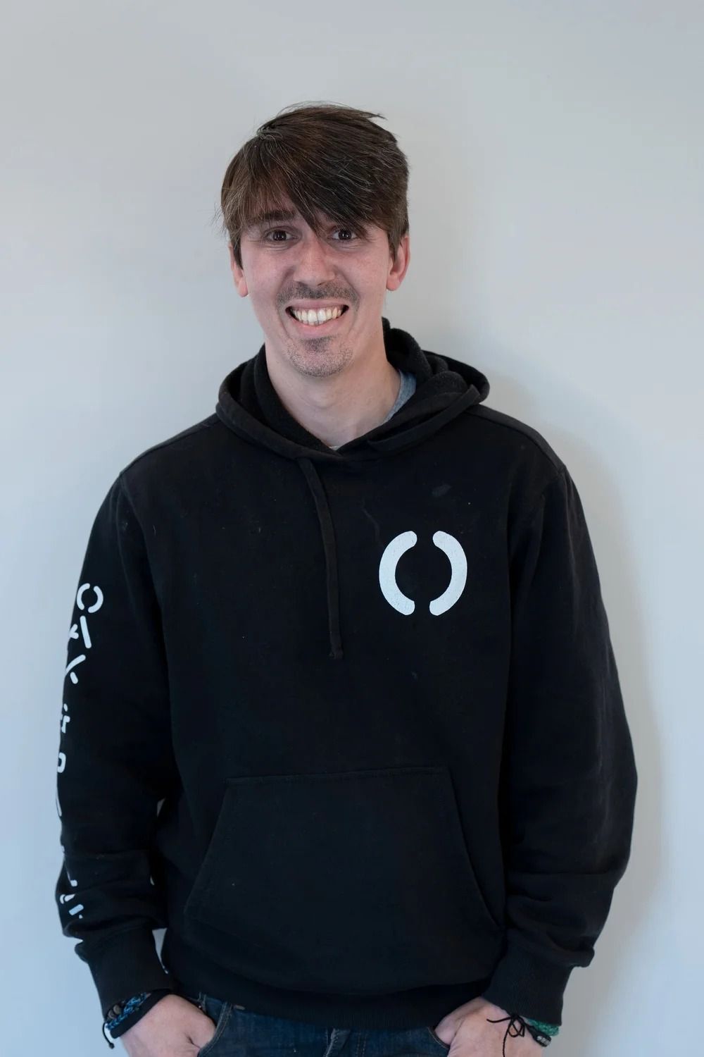 Man with brown hair wearing a black hoodie with white logo, smiling, hands in pockets.