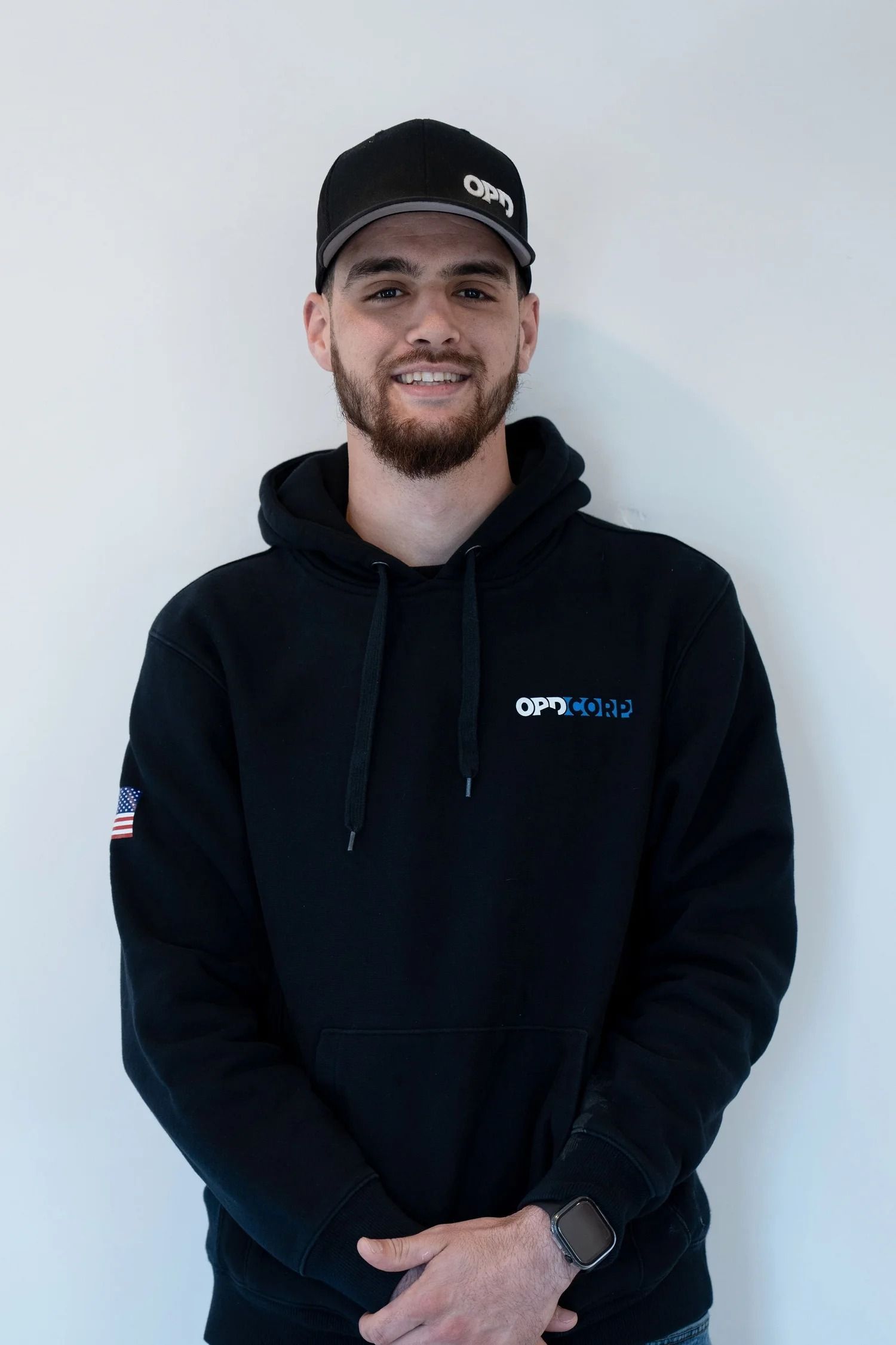 Man wearing a black hoodie and cap smiles, arms crossed, standing against a white wall.