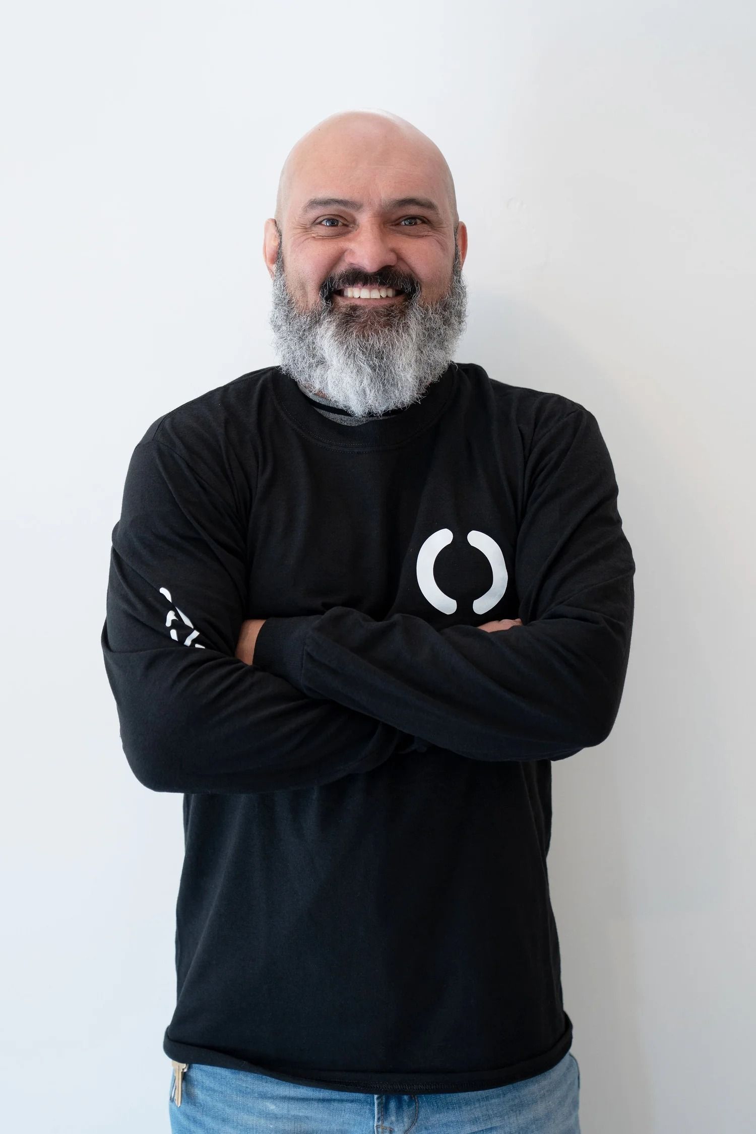 Bald man with a white beard, wearing a black long-sleeve shirt with a logo, arms crossed, smiling.