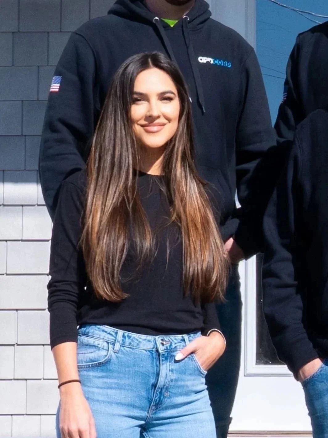 Woman in black top and jeans, smiling, with two people in hoodies behind her.