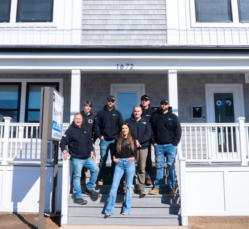 Group of people in front of a two-story building. All are wearing black hoodies and jeans. Building has white siding and gray shingles.