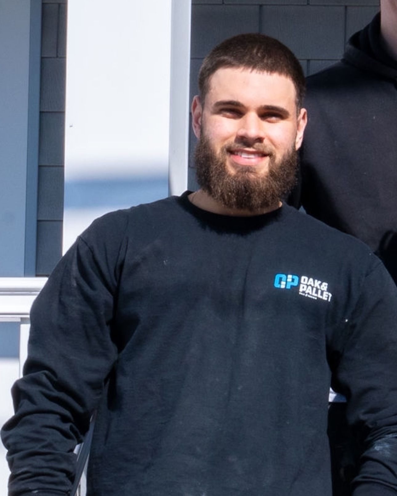 Man with beard, wearing a dark blue long-sleeved shirt with a logo, smiles outdoors.