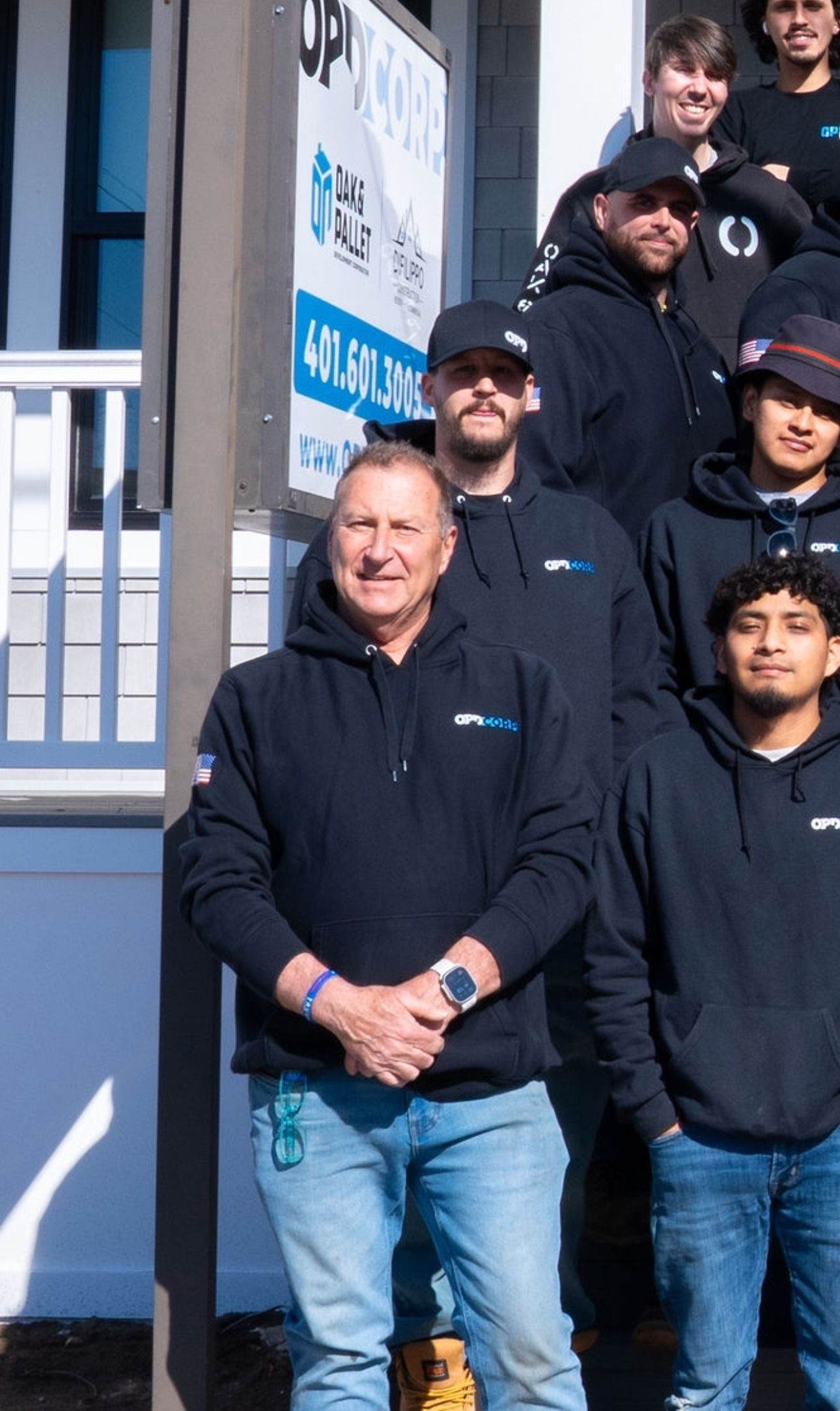 A group of men in black hoodies and jeans stand outside a building. One man is in front, arms crossed.