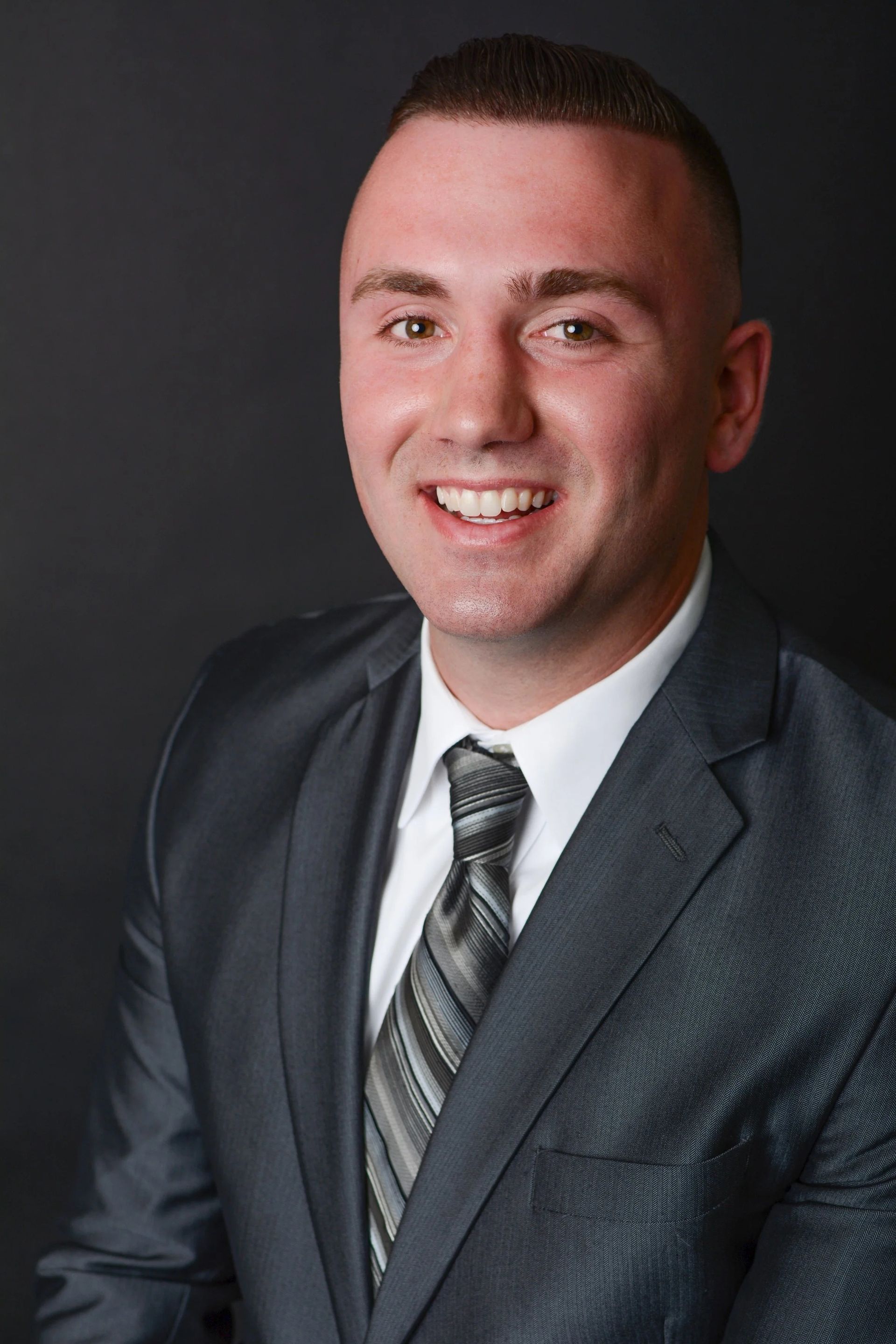 Man in a gray suit and tie smiles at the camera against a black background.