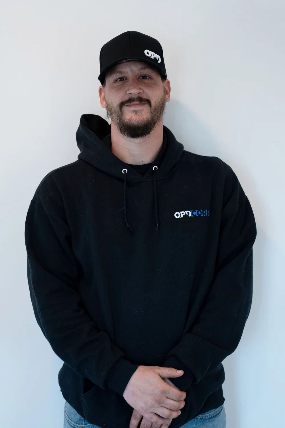 Man wearing a black hoodie and cap with logo, standing against a white wall.