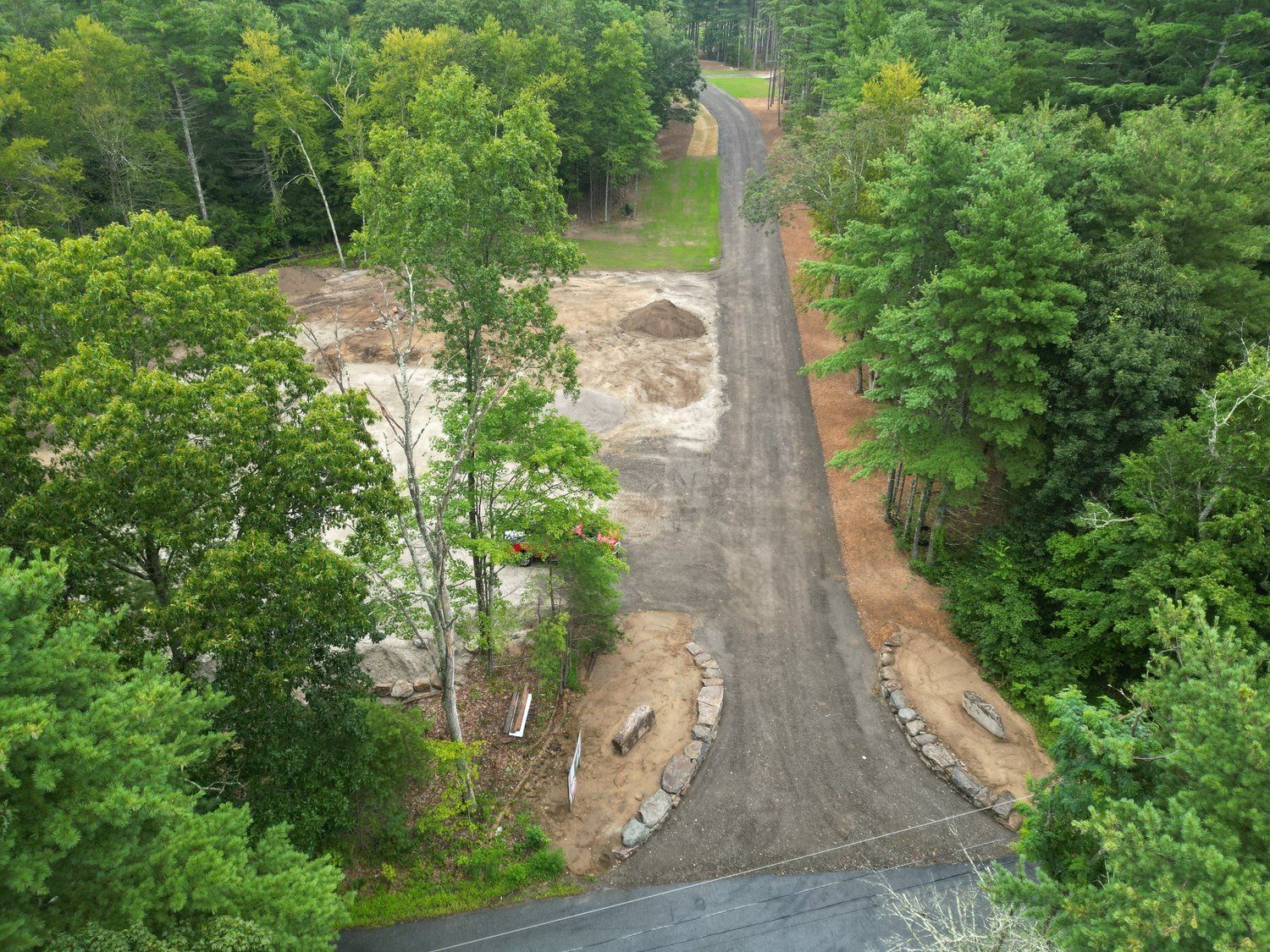 Gravel driveway leading through a forest to a cleared area with a dirt pile.