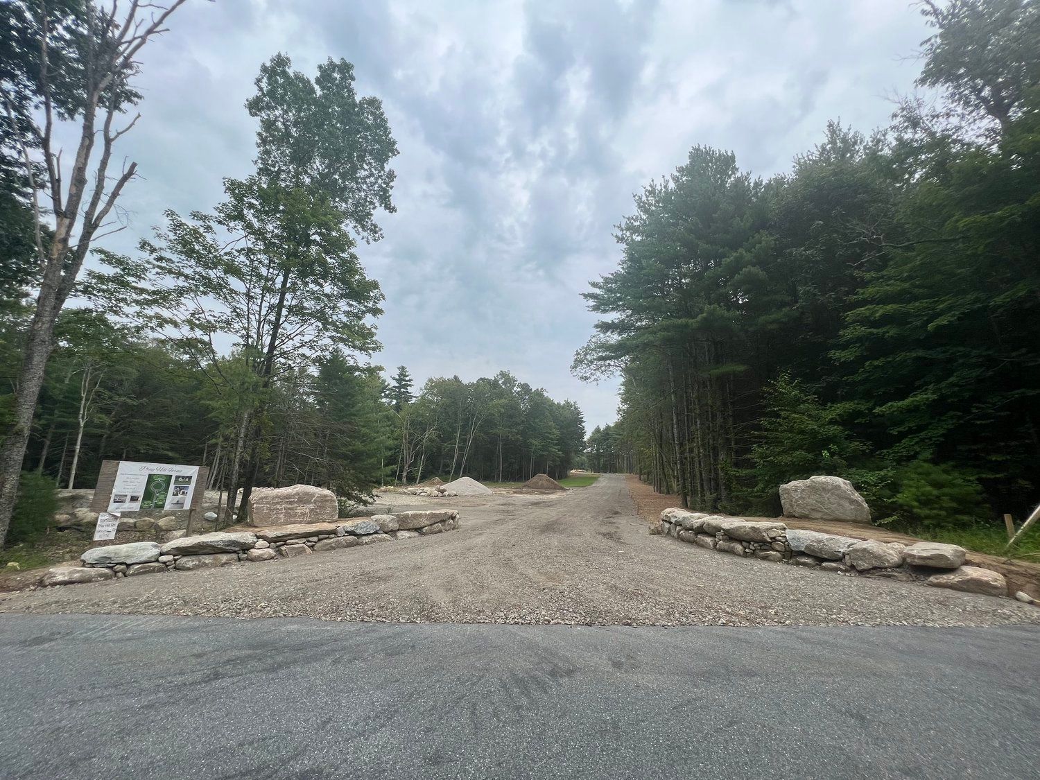 Gravel road entrance to wooded area, with stone accents and informational sign under cloudy sky.