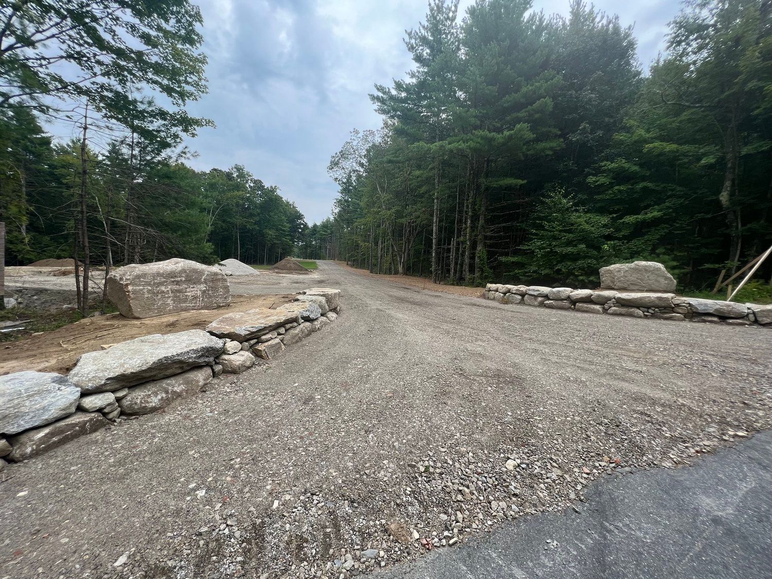 Gravel driveway with stone walls, leading into a wooded area under a cloudy sky.