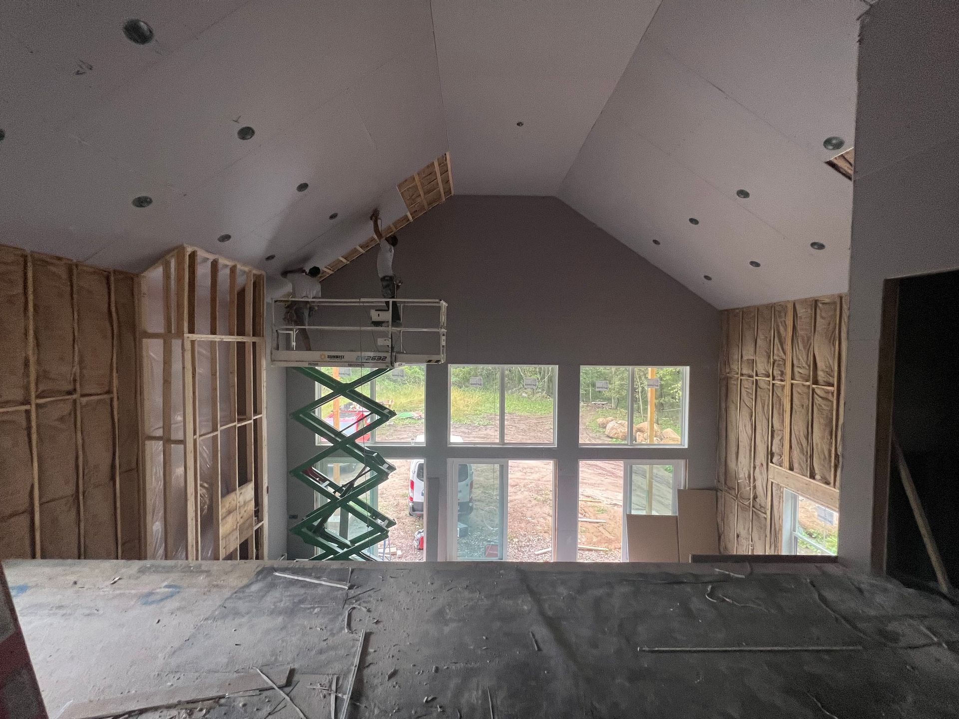Interior construction of a building with a scissor lift near large windows; wood framing and insulation visible.