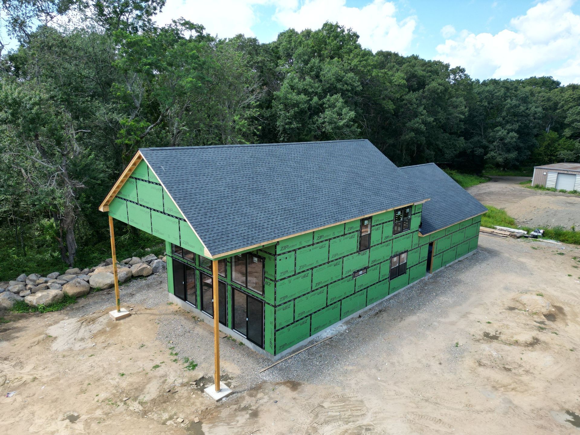 Building under construction, green sheathing, dark roof, wooden supports, gravel ground, forest background.