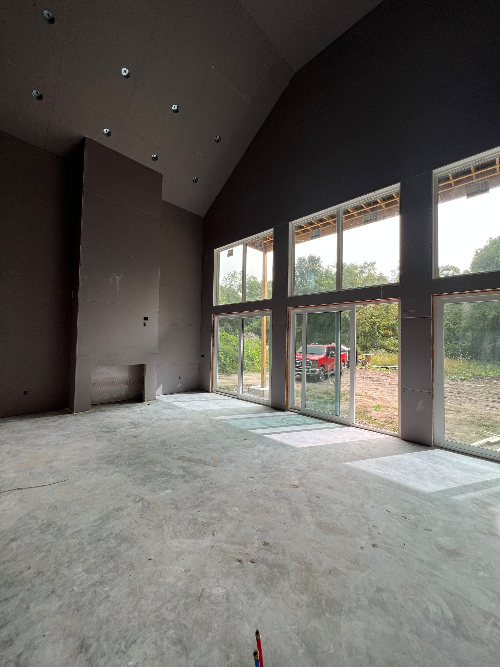 Interior of a room with large windows, unfinished walls, and concrete floor; a red truck is visible outside.