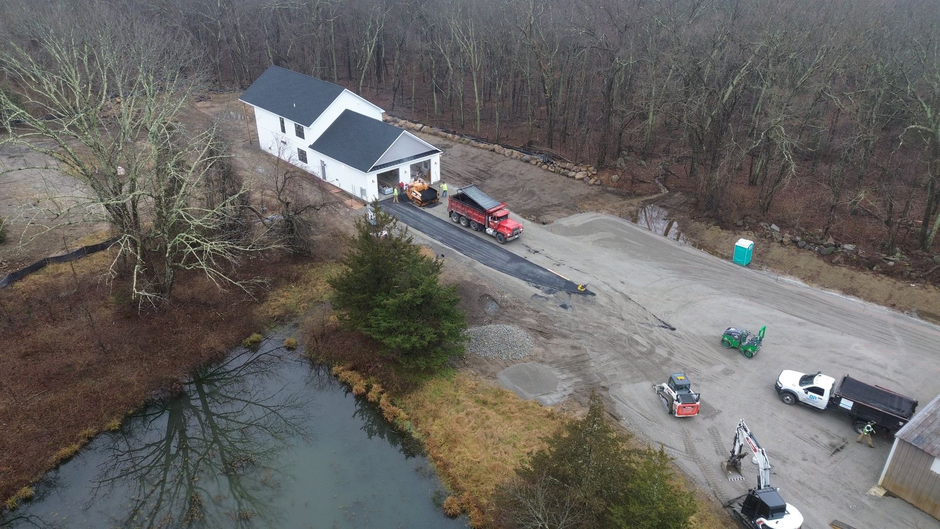 Construction site with a building, trucks, and heavy machinery near a pond and woods.