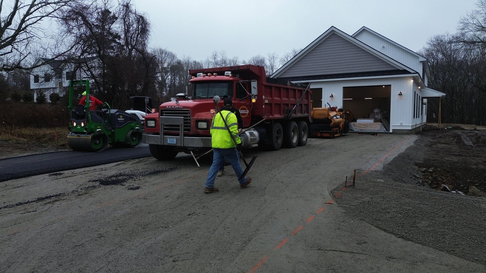 Road paving: Workers and machinery laying asphalt driveway near a house. Red dump truck, roller, and worker with compactor.