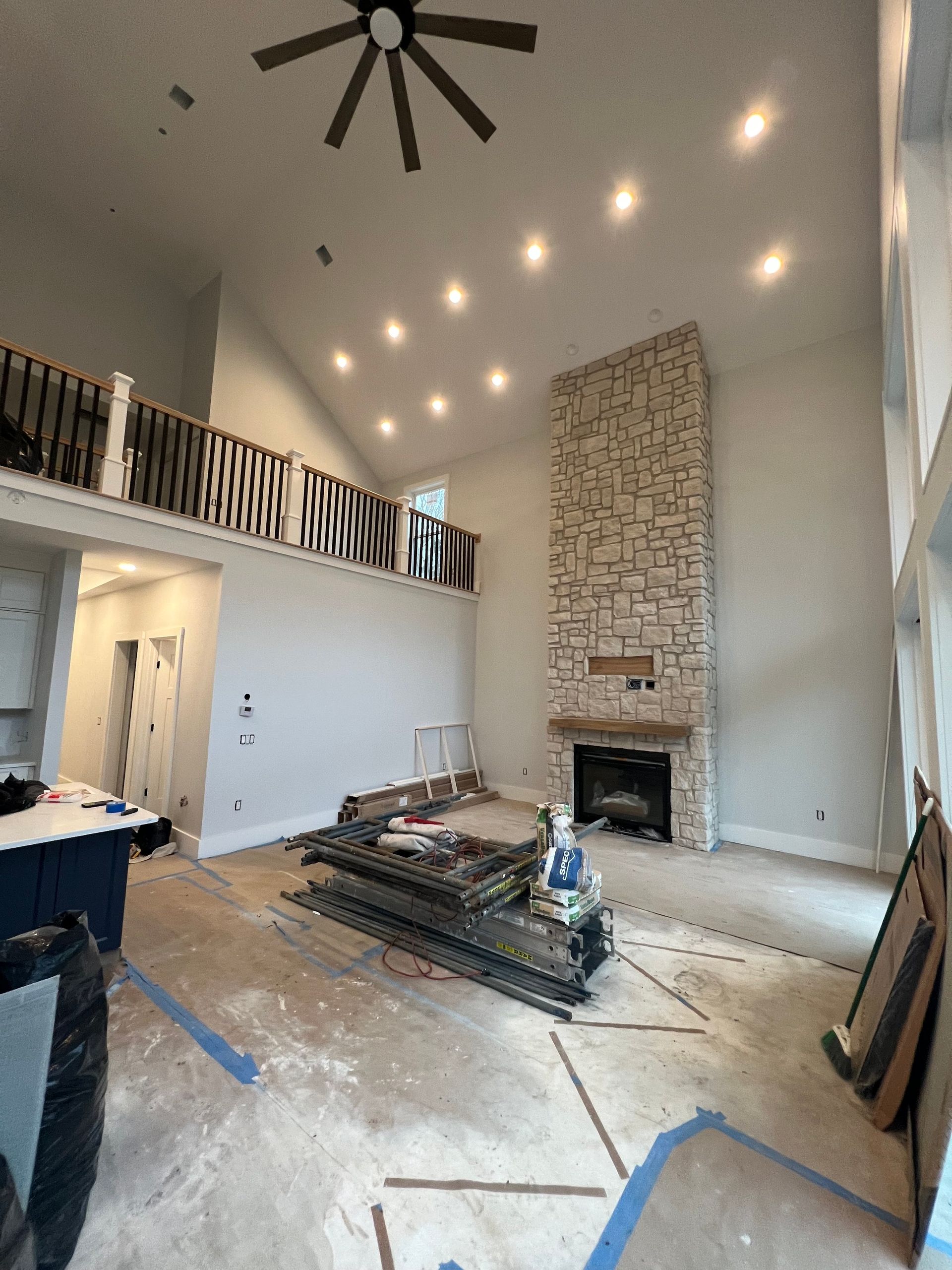 Interior of a living room under renovation with a high ceiling, fireplace, and balcony. Wood floor is being replaced.