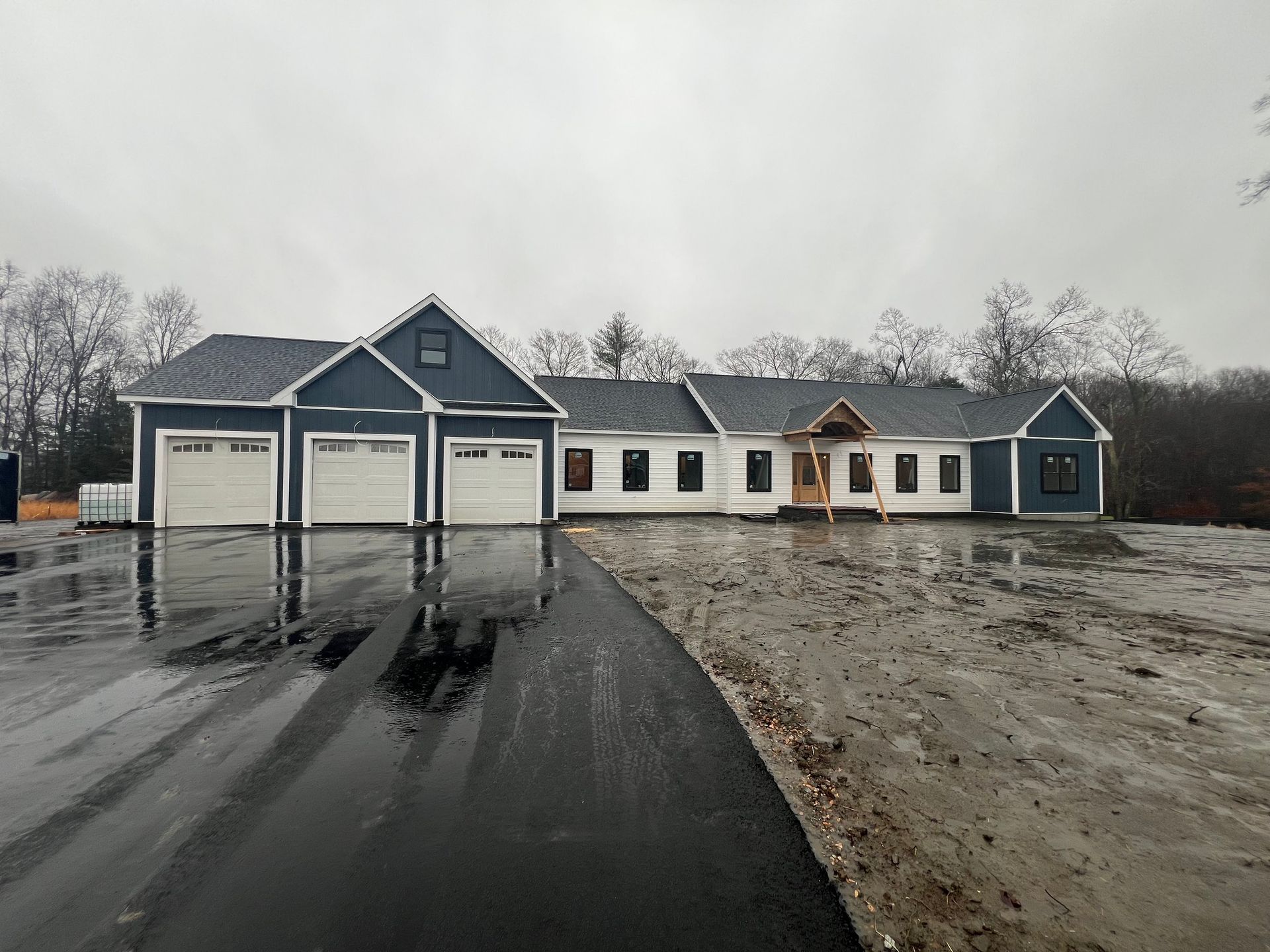 New, unfinished ranch home with three-car garage; blue and white exterior, asphalt driveway, overcast sky.