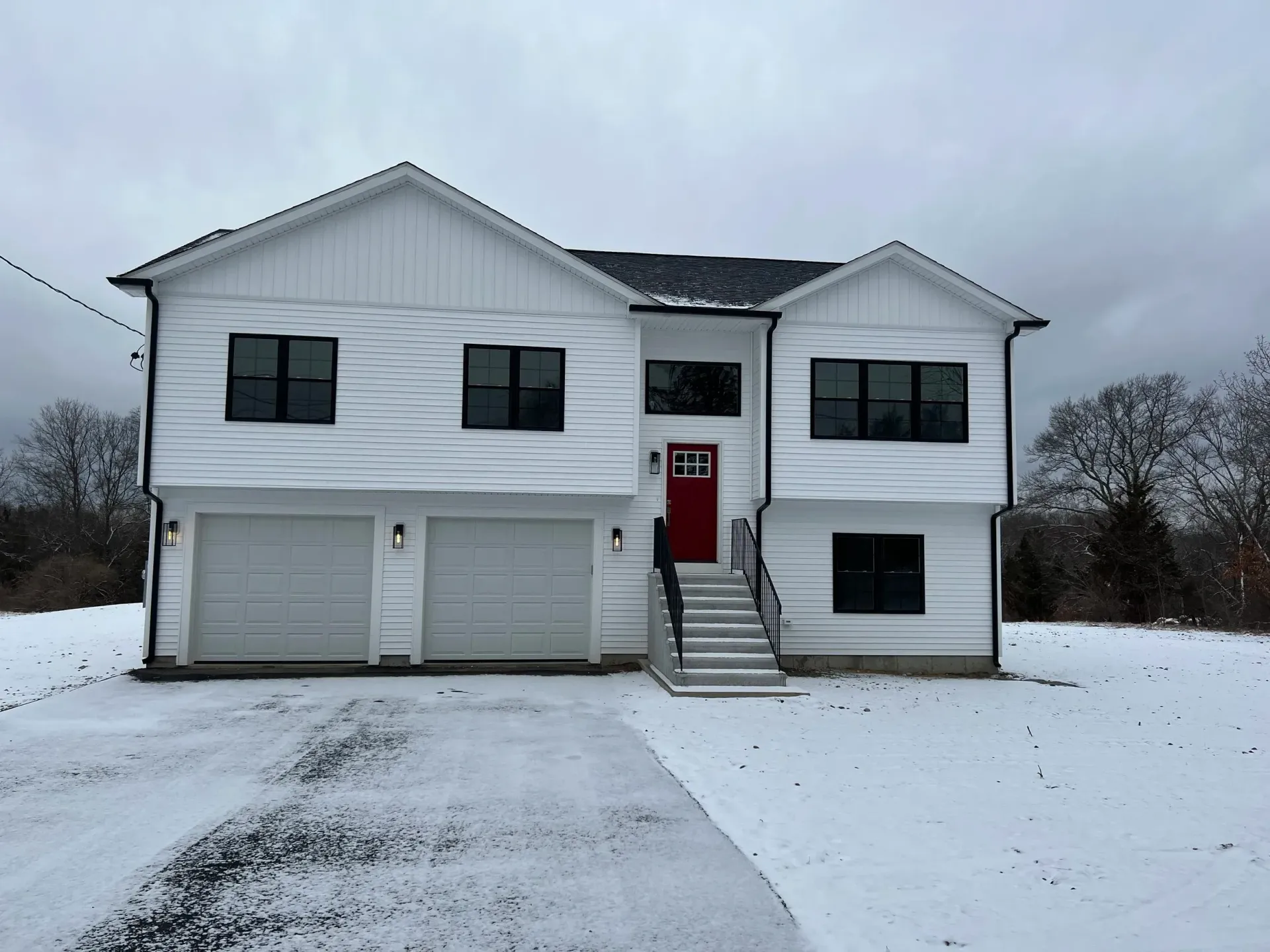 White two-story house with black windows, red front door, and two-car garage. Snow covers the ground and driveway.