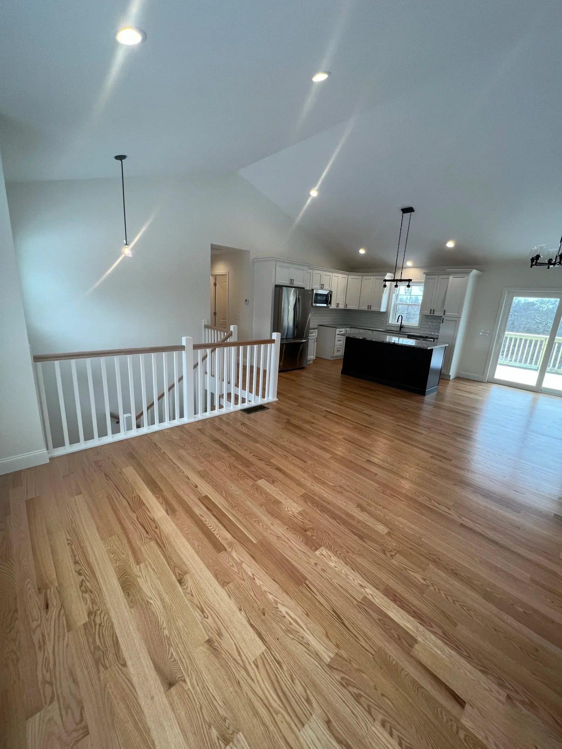 Interior view of a house with wooden floors, white walls, and a kitchen with black island. Staircase with white railing.