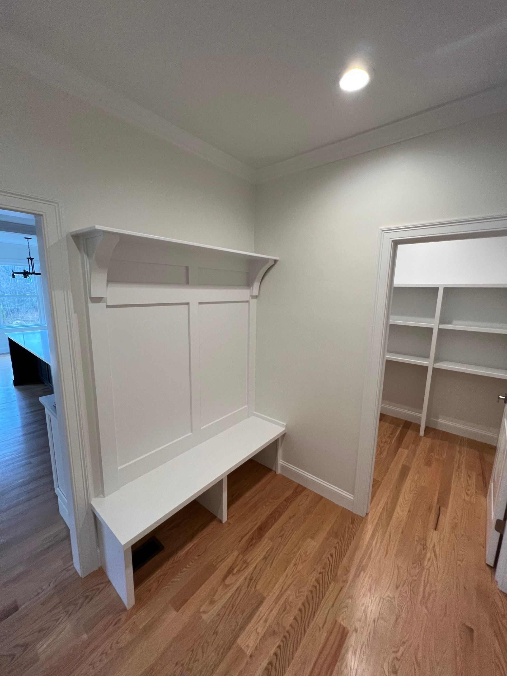 White built-in bench with shelf and coat hooks in a hallway, adjacent to a closet with shelving.