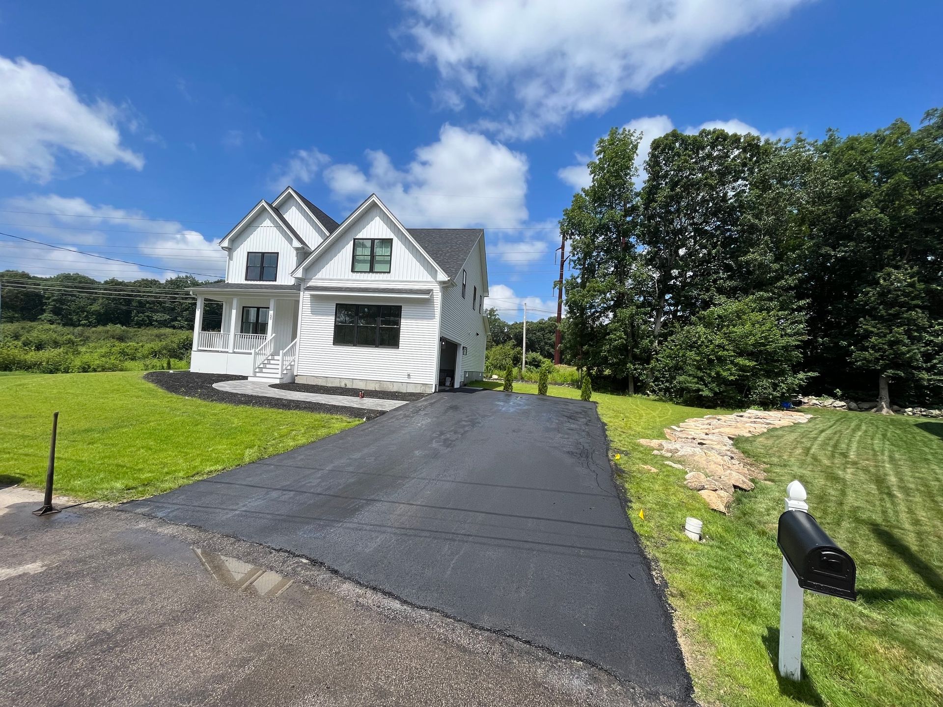 White house with new asphalt driveway on a sunny day. Green lawn and trees surround.