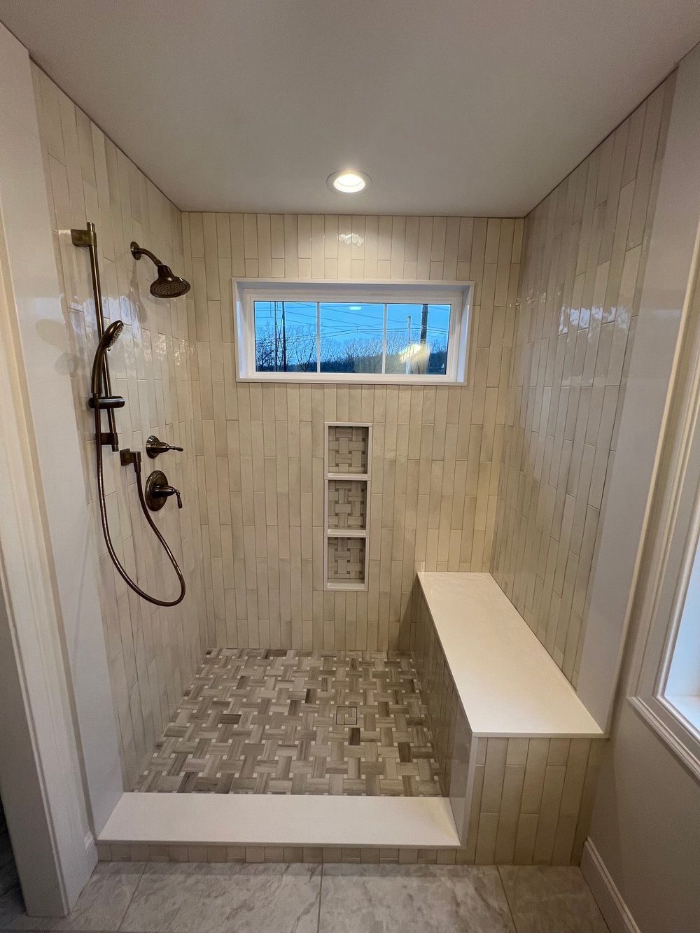 Bathroom shower with tan, vertical tile, pebble floor, built-in bench, window, and dark fixtures.
