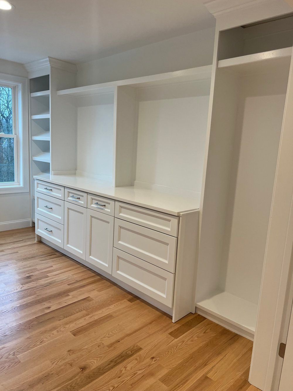 White built-in cabinetry with drawers, shelves, and open spaces in a room with hardwood floors and a window.