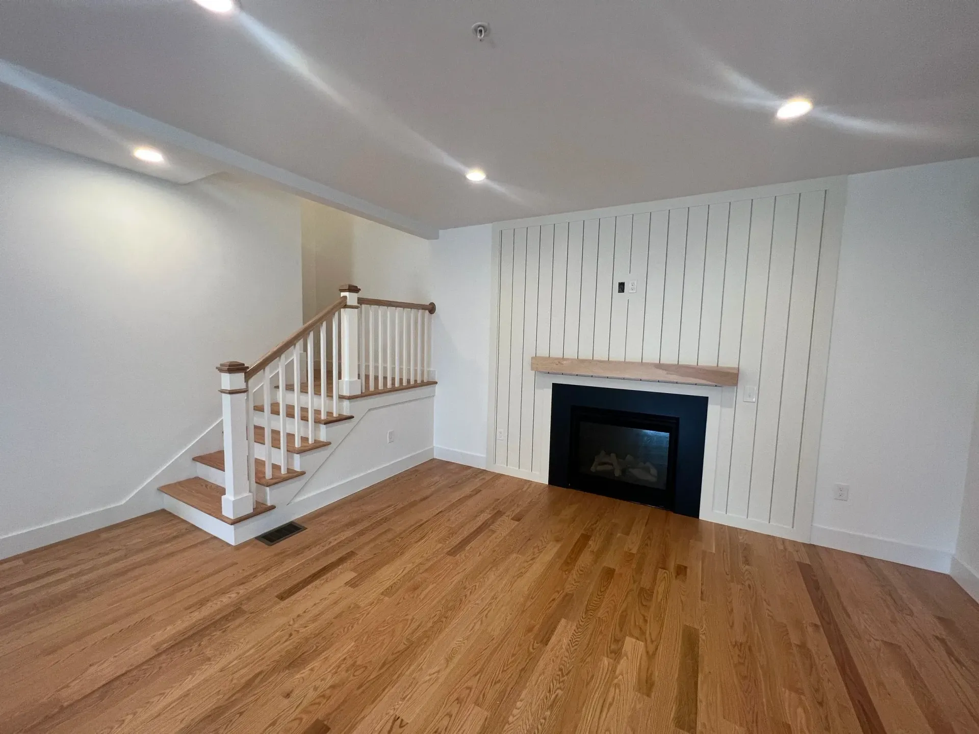 Empty living room with wood floors, fireplace, and staircase. White walls, wood trim.