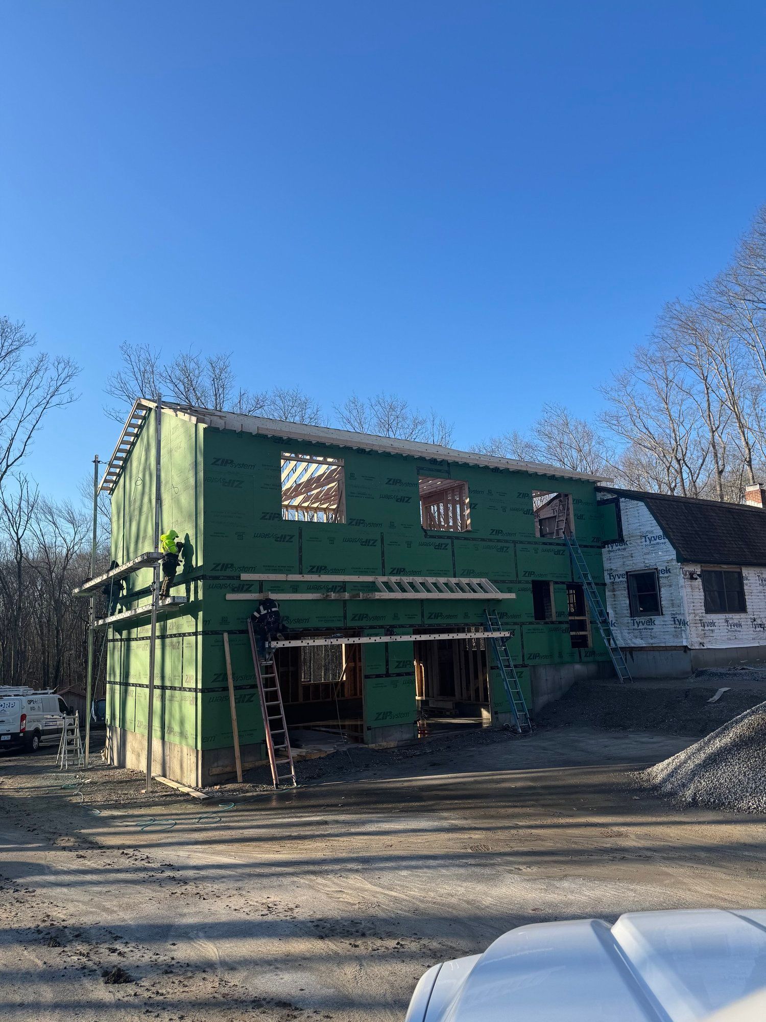 Construction site of a two-story building addition. Green sheathing, workers on scaffolding, blue sky.