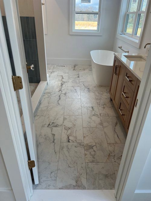 Bathroom with marble-look floor tiles, wooden vanity, and a freestanding tub near a window.