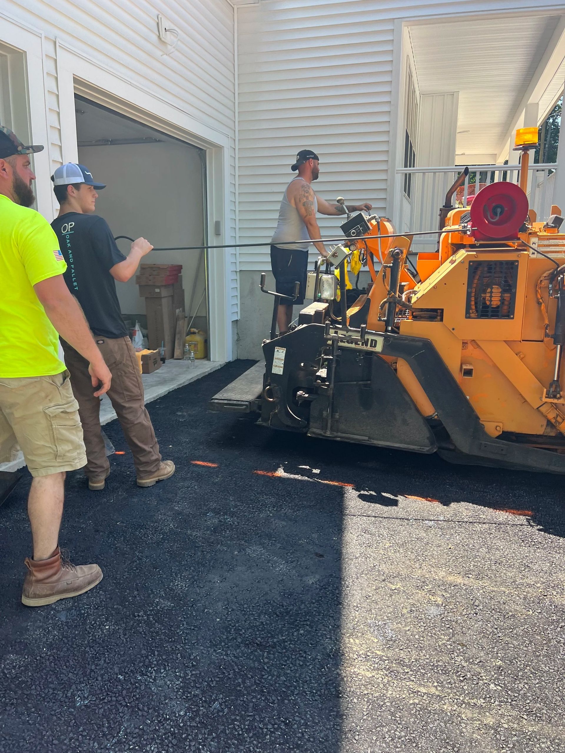 Workers paving a driveway; one operating machine, two pulling a line. Dark asphalt, bright sun.