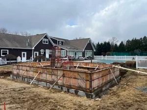 Construction site: wooden foundation formwork in front of a large house with cloudy sky.