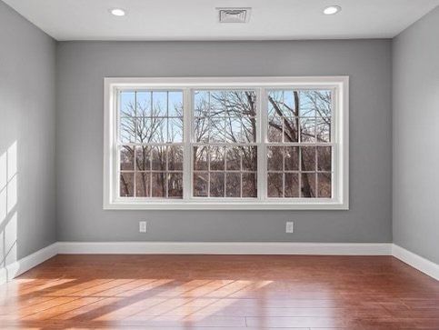 Empty room with a large window. Gray walls, wooden floor, white trim. Outside view: trees.