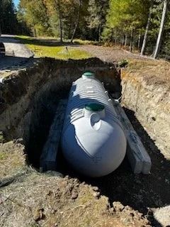 Large, gray septic tank in an excavation pit, ready for burial. Green access lids. Surrounded by dirt and grassy area.